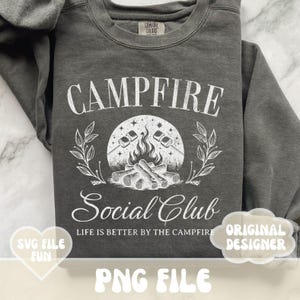 Może przedstawiać: Szara bluza z białym nadrukiem przedstawiającym ognisko z piankami i tekstem "Campfire Social Club" oraz "Life is better by the campfire".