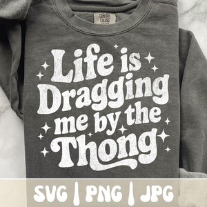 May include: A dark gray sweatshirt with the phrase "Life is Dragging me by the Thong" in white, retro-style lettering. The sweatshirt also has the words "SVG | PNG | JPG" at the bottom.