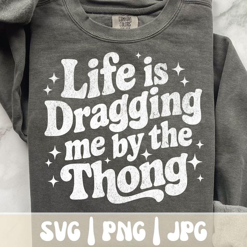 May include: A dark gray sweatshirt with the phrase "Life is Dragging me by the Thong" in white, retro-style lettering. The sweatshirt also has the words "SVG | PNG | JPG" at the bottom.