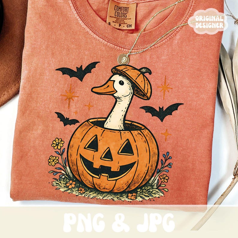 May include: A coral-colored t-shirt featuring a Halloween-themed graphic. The design includes a white goose wearing a pumpkin hat, emerging from a carved pumpkin. Black bats and stars surround the pumpkin. The shirt has the text "PNG & JPG".
