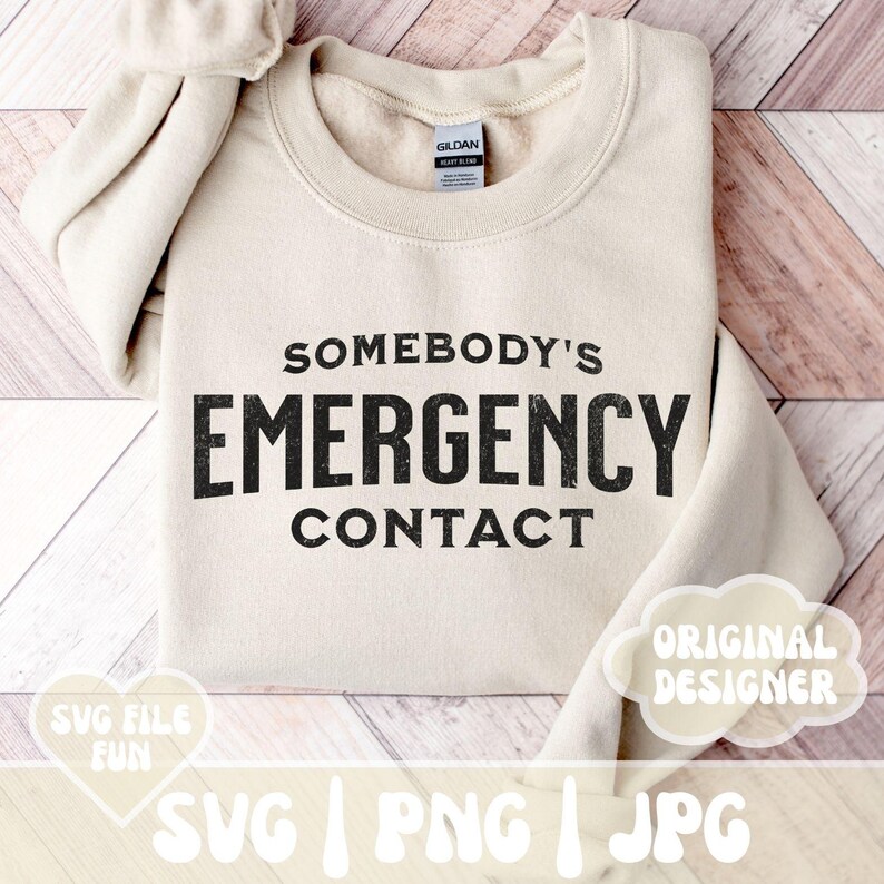 以下が含まれることがあります： 木製の表面に置かれた、ベージュのトレーナー。トレーナーには「Somebody's Emergency Contact」という黒い文字が、かすれたような書体で書かれている。