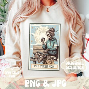 K&ouml;nnte beinhalten: Eine digitale Illustration eines Skeletts, das auf einer Bank mit einer Tasse Kaffee sitzt. Das Skelett tr&auml;gt ein blaues Hoodie mit einem Peace-Zeichen. Der Text "The Tired Mom" befindet sich unter dem Bild.