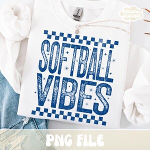May include: White sweatshirt with the text "SOFTBALL VIBES" in blue, featuring a checkered pattern and star accents. The sweatshirt is folded, with a necklace visible. The words "PNG FILE" are at the bottom.