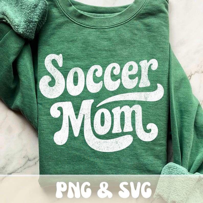 Op de afbeelding: Een groene sweatshirt met de tekst "Soccer Mom" in grote, witte letters in retrostijl. De sweatshirt is opgevouwen, de mouwen zijn zichtbaar. De tekst "PNG & SVG" staat onderaan.