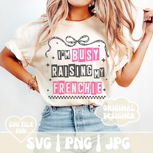 K&ouml;nnte beinhalten: Ein wei&szlig;es T-Shirt mit schwarzem Text, der "I'm Busy Raising My Frenchie" lautet. Der Text ist in einer fetten, klobigen Schriftart gehalten und mit Pink umrandet. Das Shirt wird von einer Person mit langen blonden Haaren getragen und zu einer blauen Jeans kombiniert.
