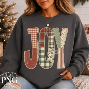 May include: A charcoal grey long-sleeve sweatshirt featuring the word JOY in large, block-style letters. Each letter is filled with a different patchwork pattern, including red and white gingham, red and green plaid, and floral prints.