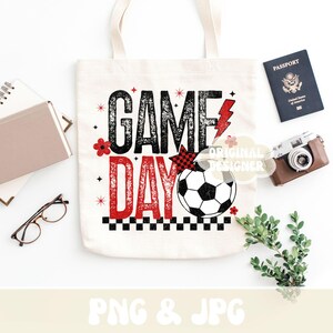 May include: A white tote bag with the words "GAME DAY" in black and red, featuring a soccer ball graphic. The bag is surrounded by a passport, camera, glasses, and greenery, suggesting a travel or sports theme.