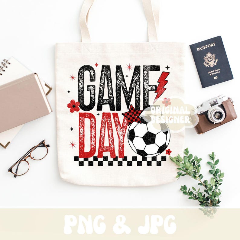 May include: A white tote bag with the words "GAME DAY" in black and red, featuring a soccer ball graphic. The bag is surrounded by a passport, camera, glasses, and greenery, suggesting a travel or sports theme.