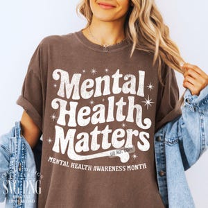 May include: A brown t-shirt with the words "Mental Health Matters" in a white, retro font. The shirt also includes the text "Mental Health Awareness Month." The design is surrounded by starbursts.