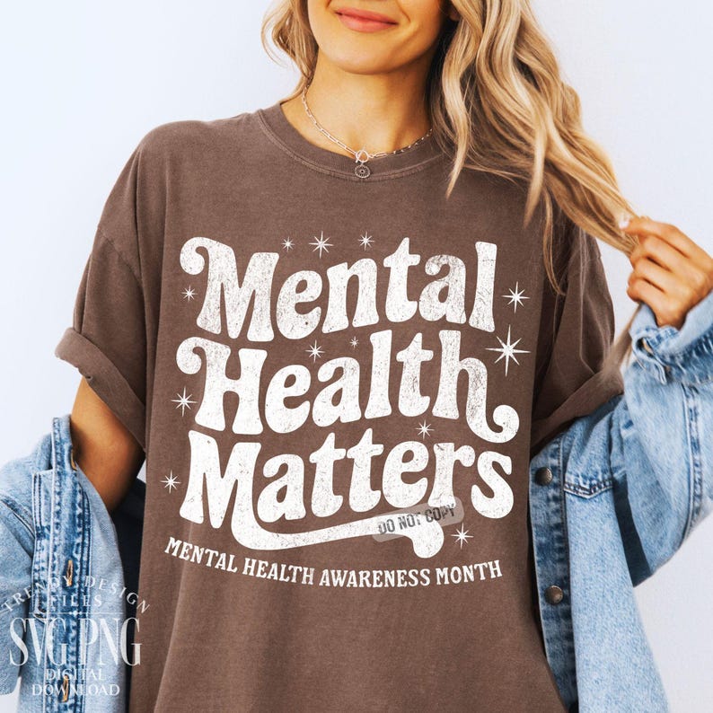 May include: A brown t-shirt with the words "Mental Health Matters" in a white, retro font. The shirt also includes the text "Mental Health Awareness Month." The design is surrounded by starbursts.