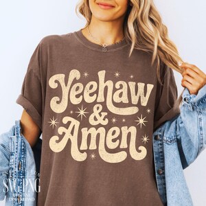 Puede incluir: Camiseta marr&oacute;n con el texto "Yeehaw & Amen" en estilo retro. El texto es de color beige claro con detalles de estrellas. La camiseta tiene mangas enrolladas y un corte casual.