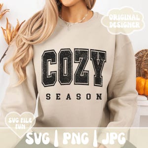 May include: A beige sweatshirt with the text "Cozy Season" in black with a distressed, vintage look. The text is in a large, bold font.