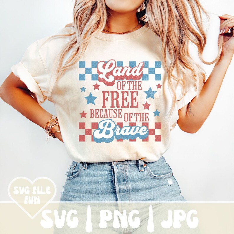 May include: Cream-colored t-shirt with the text "Land of the Free Because of the Brave" in red, white, and blue. The design includes stars and checkered patterns, evoking a patriotic theme. The shirt is made of a soft material.