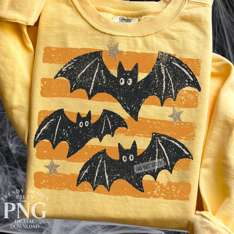 May include: A pale yellow long-sleeve shirt featuring a graphic print of three black bats with white eyes, positioned over horizontal orange distressed stripes and small grey stars. The inner collar tag displays the text Comfort Colors.