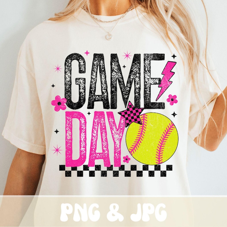 May include: Cream-colored t-shirt with the words "GAME DAY" in black and pink, featuring a softball graphic. The design includes a pink lightning bolt, a checkered star, and floral accents. The bottom of the shirt has a checkered border.