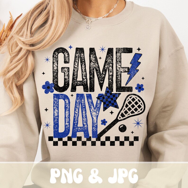May include: A beige sweatshirt with the words "GAME DAY" in black and blue, with a lacrosse stick graphic. The design includes a lightning bolt, stars, and flowers. The bottom of the design has a black and white checkered pattern.