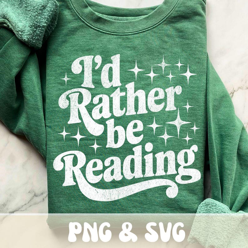 Op de afbeelding: Groen sweatshirt met de witte tekst "I'd Rather be Reading", met steraccenten. De onderkant van de trui heeft de witte tekst "PNG & SVG". De trui is op een wit marmeren oppervlak gevouwen.