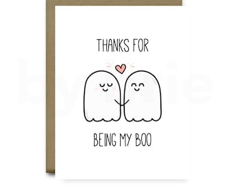 May include: A white card with a brown envelope. The card has a black and white illustration of two ghosts holding hands with a pink heart above them. The text on the card reads "Thanks for being my boo."