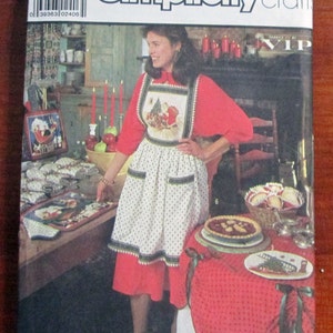 May include: A Simplicity Crafts sewing pattern envelope for Christmas table accessories, a tote bag, and an apron. The cover features a photograph of a person wearing a white apron with a Santa Claus graphic over a red long-sleeve shirt and skirt.