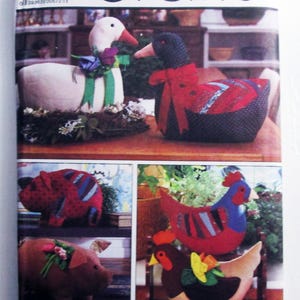 May include: A Simplicity Crafts sewing pattern envelope 5148 by Marjorie Puckett featuring illustrations of stuffed animal geese, pigs, and chickens. The geese wear fabric bows, the pigs have floral accents, and the chickens are multi-colored.