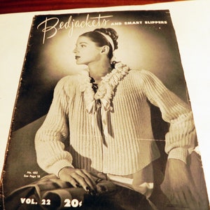 May include: A vintage magazine cover titled Bedjackets and Smart Slippers Vol. 22, featuring a black and white photograph of a person wearing a light-colored, ribbed knit bedjacket with a ruffled collar and ribbon tie, No. 485, 20 cents.