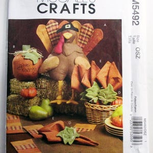 May include: McCall's Crafts M5492 sewing pattern envelope featuring a fabric turkey, a pumpkin with the text give thanks, fabric leaves, and table linens. The design includes plaid and solid brown fabrics, a woven basket, and pears.