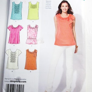 May include: Simplicity sewing pattern featuring various blouse designs with ruffled necklines. The pattern includes sizes 12-20 and is suitable for B, C, and D cup sizes. The image shows a woman wearing a coral-colored blouse with a ruffled neckline.