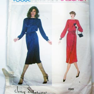May include: Vintage Vogue American Designer sewing pattern featuring two dress designs. One dress is a long-sleeved, navy blue dress with a belt. The other is a long-sleeved, red dress with a button-up front and a belt. The pattern is signed by Jerry Silverman.