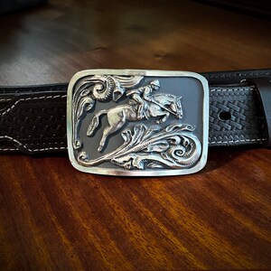May include: A silver belt buckle with an intricate design of a horse and rider. The buckle is set against a dark gray background and features a floral motif.