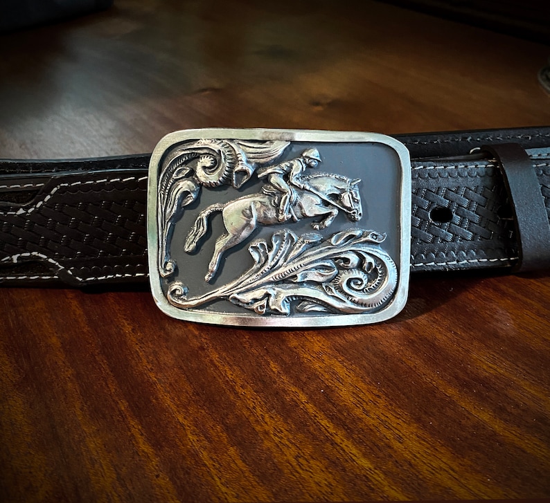 May include: A silver belt buckle with an intricate design of a horse and rider. The buckle is set against a dark gray background and features a floral motif.