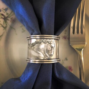 May include: Silver napkin ring with a detailed engraved image of a horse's head. The ring is holding a dark blue napkin.
