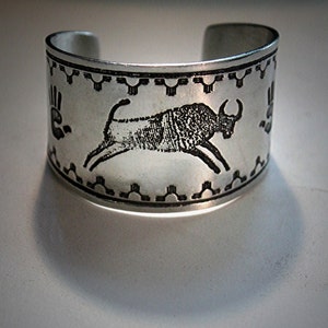 May include: A silver metal bracelet featuring a black and white image of a bull running. The bracelet has a decorative border with a repeating pattern.