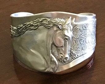 May include: A silver bracelet with a detailed horse head design. The horse's mane is flowing and the design is intricate. The bracelet is curved and has a smooth finish.
