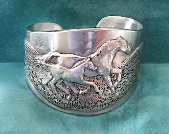 May include: A silver bracelet with a detailed design of three horses running. The horses are in a raised relief, and the background is textured.