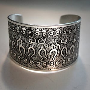 May include: A silver cuff bracelet with an etched design of stylized figures dancing. The figures are repeated around the entire bracelet.