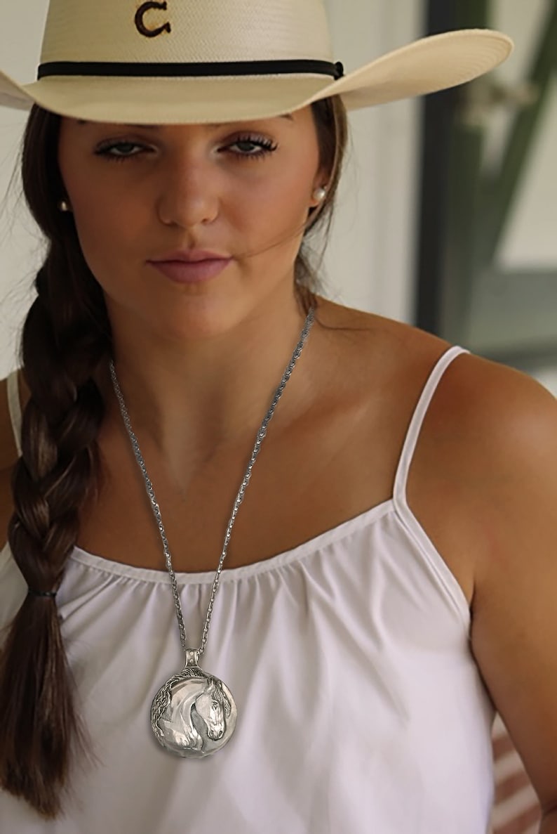 May include: A woman wearing a white tank top and a straw cowboy hat with a black band. She is wearing a silver necklace with a horse head pendant.