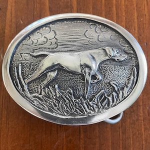 May include: Silver belt buckle with a detailed design of a dog running through tall grass. The dog is in a hunting pose with its head turned to the side. The background is a cloudy sky.