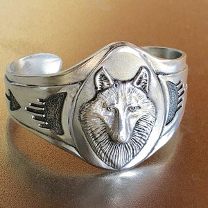 May include: A silver ring with a wolf head design. The ring is detailed with a wolf's face in the center and a Native American-inspired design on the sides.
