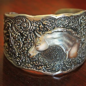May include: Silver-toned cuff bracelet with an embossed design. The bracelet features a detailed relief of a boar's head and intricate floral patterns. The metal has a weathered appearance, adding to its rustic charm.