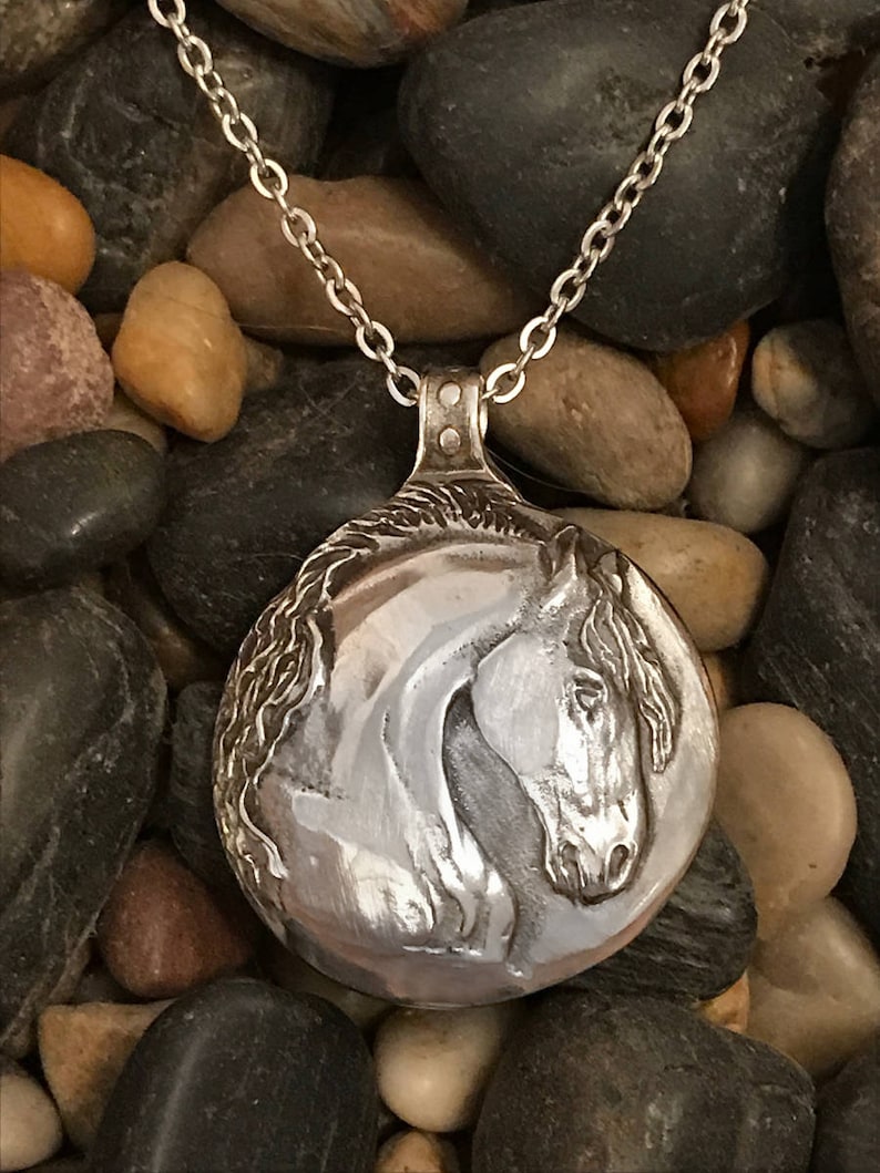 May include: A silver horse head pendant necklace on a silver chain. The pendant is round and features a detailed horse head design. The necklace is displayed on a bed of small, smooth stones.
