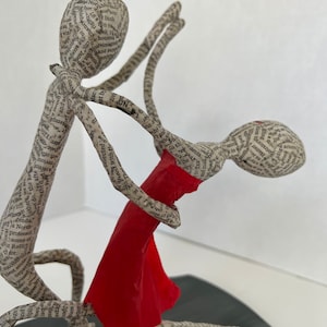 Pu&ograve; includere: Una scultura in cartapesta di due figure che ballano. La donna indossa un vestito rosso e l'uomo la tiene sollevata in aria. Le figure sono fatte di carta e sono ricoperte di testo.