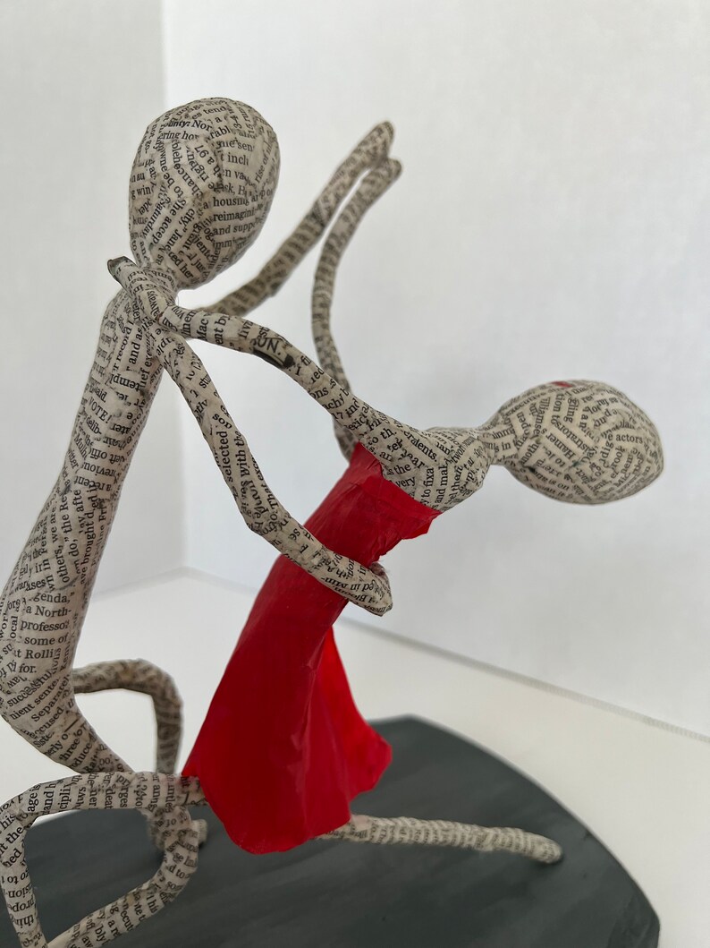Pu&ograve; includere: Una scultura in cartapesta di due figure che ballano. La donna indossa un vestito rosso e l'uomo la tiene sollevata in aria. Le figure sono fatte di carta e sono ricoperte di testo.