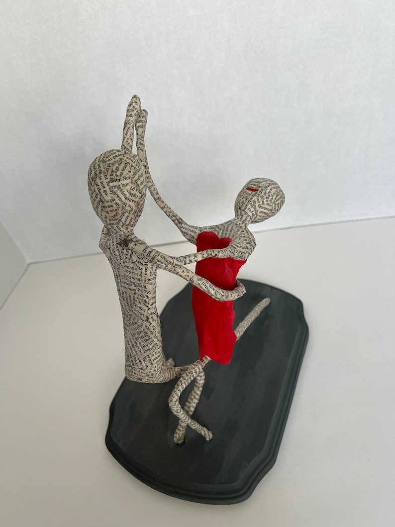 Pu&ograve; includere: Una scultura in cartapesta di una coppia che balla. L'uomo indossa una camicia bianca e la donna un vestito rosso. La scultura &egrave; su una base nera.