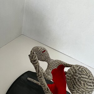 Pu&ograve; includere: Una scultura di due figure che si abbracciano, realizzata in carta e ricoperta di testo. La figura a sinistra indossa un vestito rosso. La scultura &egrave; su una base nera.