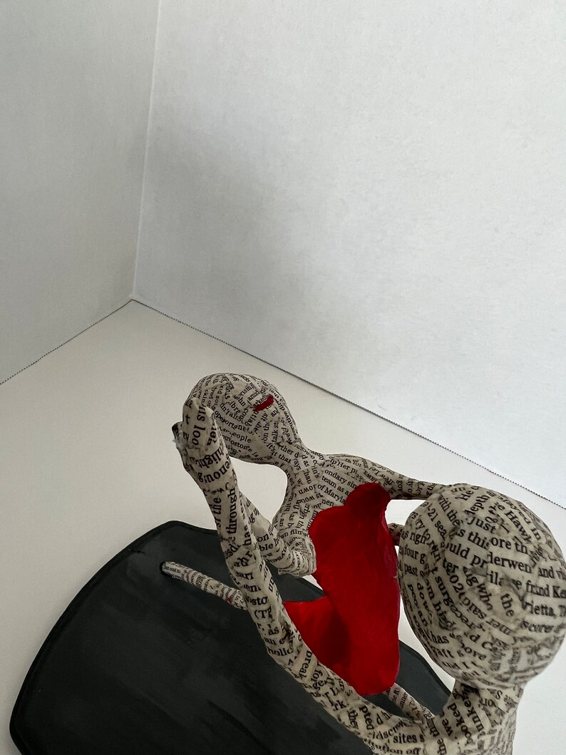 Pu&ograve; includere: Una scultura di due figure che si abbracciano, realizzata in carta e ricoperta di testo. La figura a sinistra indossa un vestito rosso. La scultura &egrave; su una base nera.