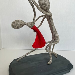 Pu&ograve; includere: Una scultura in cartapesta di una coppia che balla. L'uomo tiene la donna tra le braccia. Entrambi sono fatti di carta e la donna indossa un vestito rosso. La scultura &egrave; su una base di legno grigio scuro.