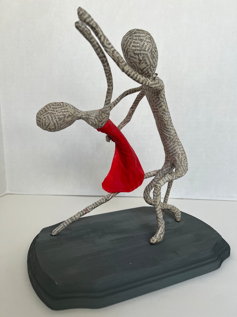 Pu&ograve; includere: Una scultura in cartapesta di una coppia che balla. L'uomo tiene la donna tra le braccia. Entrambi sono fatti di carta e la donna indossa un vestito rosso. La scultura &egrave; su una base di legno grigio scuro.