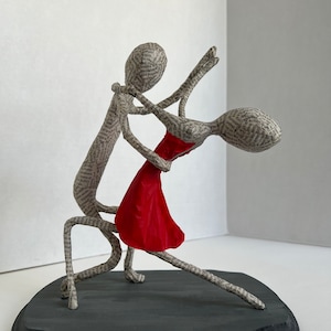 Pu&ograve; includere: Una scultura in cartapesta di una coppia che balla. L'uomo tiene la donna in una posizione di tuffo. La donna indossa un vestito rosso. La scultura &egrave; su una base di legno nero.