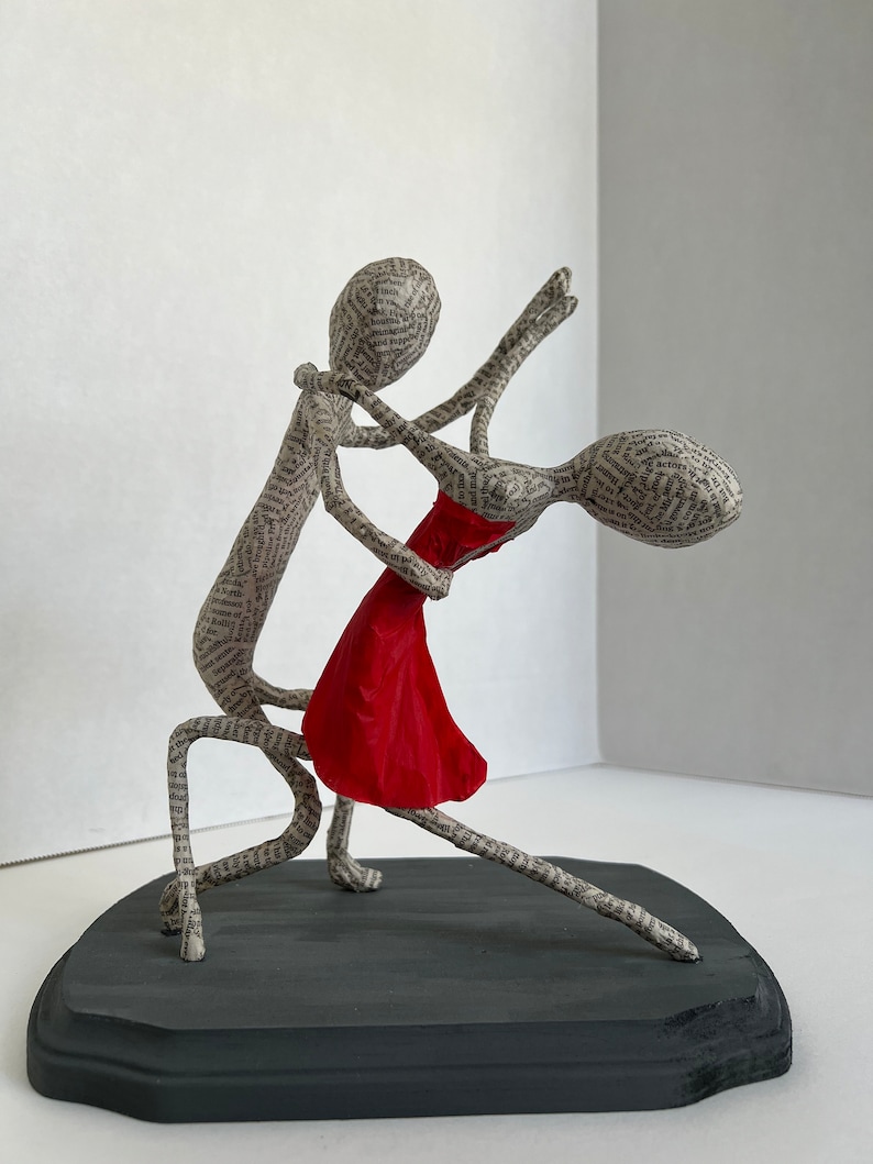 Pu&ograve; includere: Una scultura in cartapesta di una coppia che balla. L'uomo tiene la donna in una posizione di tuffo. La donna indossa un vestito rosso. La scultura &egrave; su una base di legno nero.