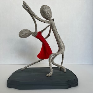 Pu&ograve; includere: Una scultura in cartapesta di una coppia che balla. L'uomo tiene la mano della donna e entrambi sorridono. La scultura &egrave; dipinta in tonalit&agrave; di grigio e rosso.